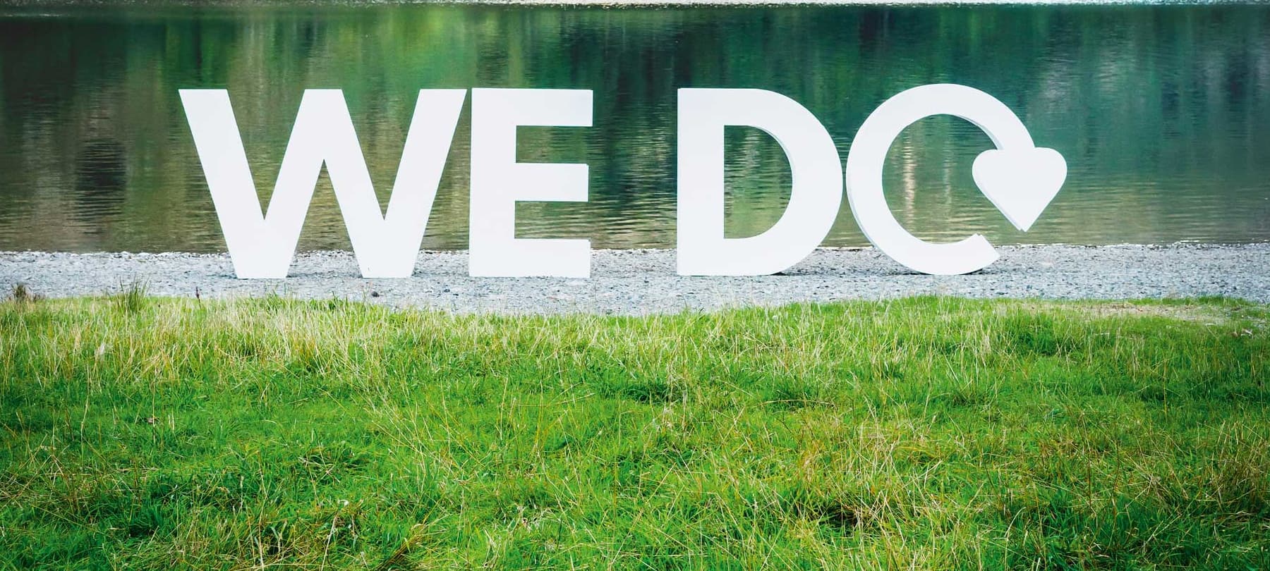 Llythrennau gwyn fawr ar lan llyn yn gwneud y geiriau "We Do". Mae'r llythr "o" wedi cael ei newid i'r logo ailgylchu